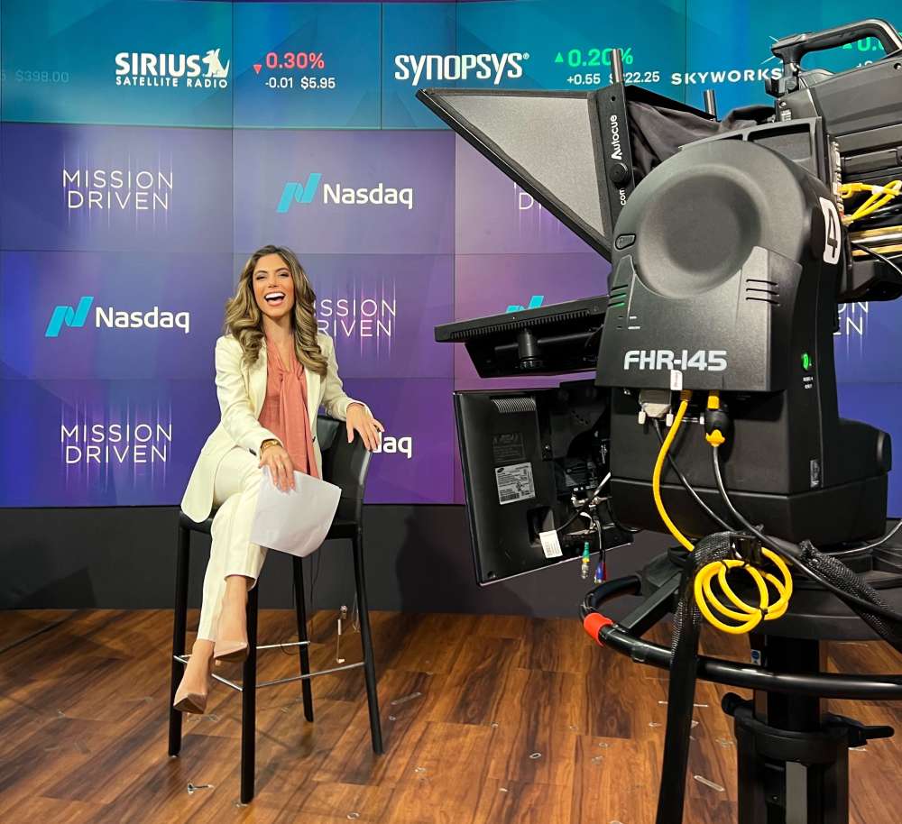 Kristina Ayanian ’19 smiles as she sits in a chair in the Nasdaq studio where she films "Mission Driven." She wears a cream colored pantsuit and rose-pink blouse. In the foreground is a television camera.
