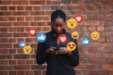 A young Black woman stands in front of a brick wall, staring down at her smartphone. A dozen different social media icons and emojis — including hearts, thumbs-up "likes" and smiley faces — surround her.