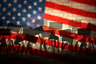 Photo illustration of brick wall with the American flag superimposed upon it and featuring shadows of raised protest placards and bullhorns.