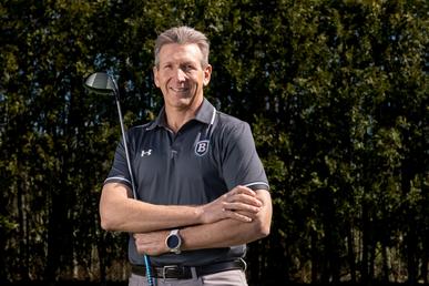 Scholnick poses with crossed arms and a golf club. He is wearing a gray polo with the Bentley logo.
