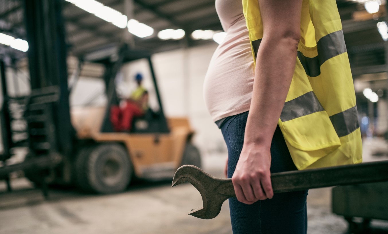 Close-up photo of pregnant construction worker's torso, with forklift in background