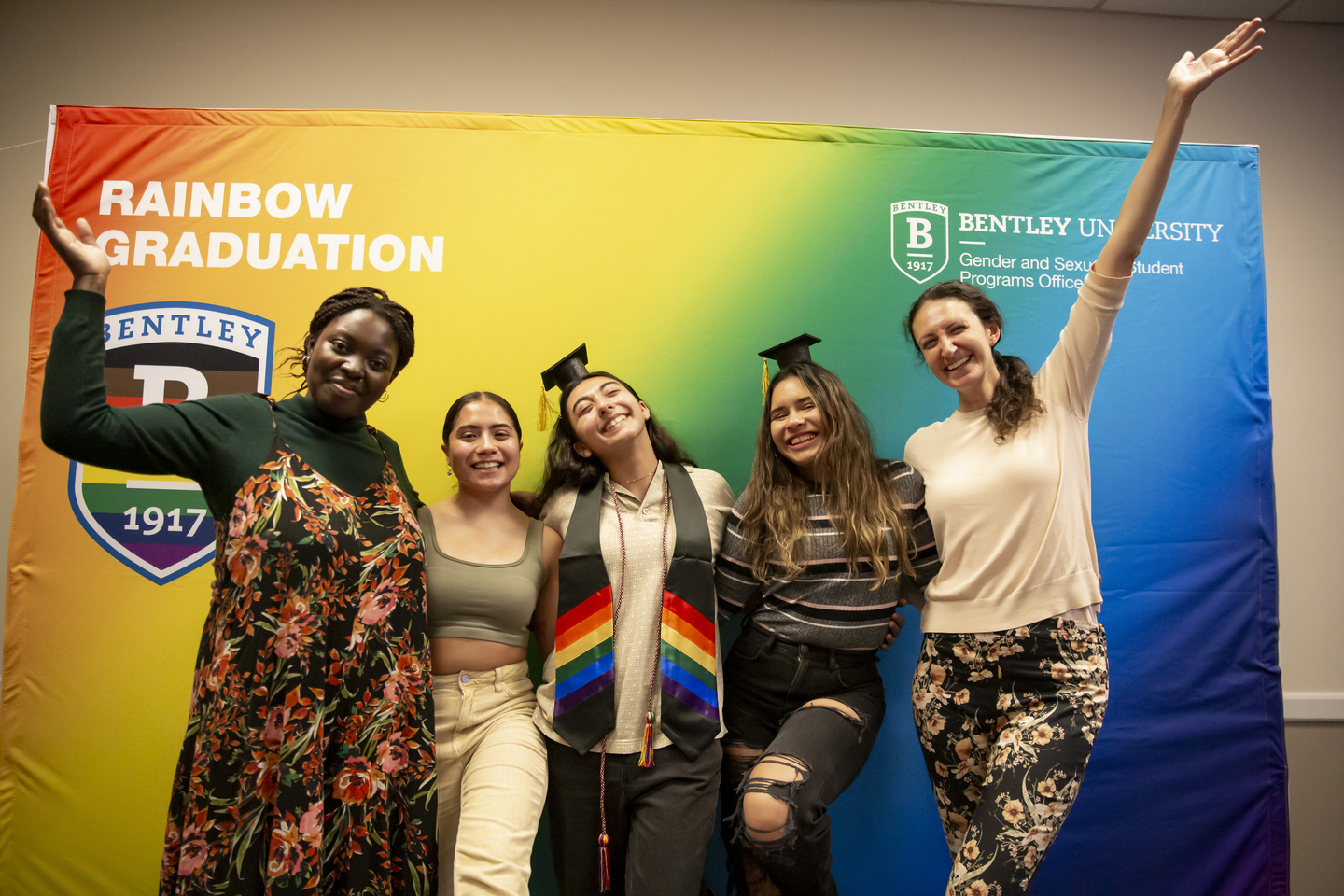 Bentley students and staff celebrate Rainbow graduation in front of rainbow backdrop