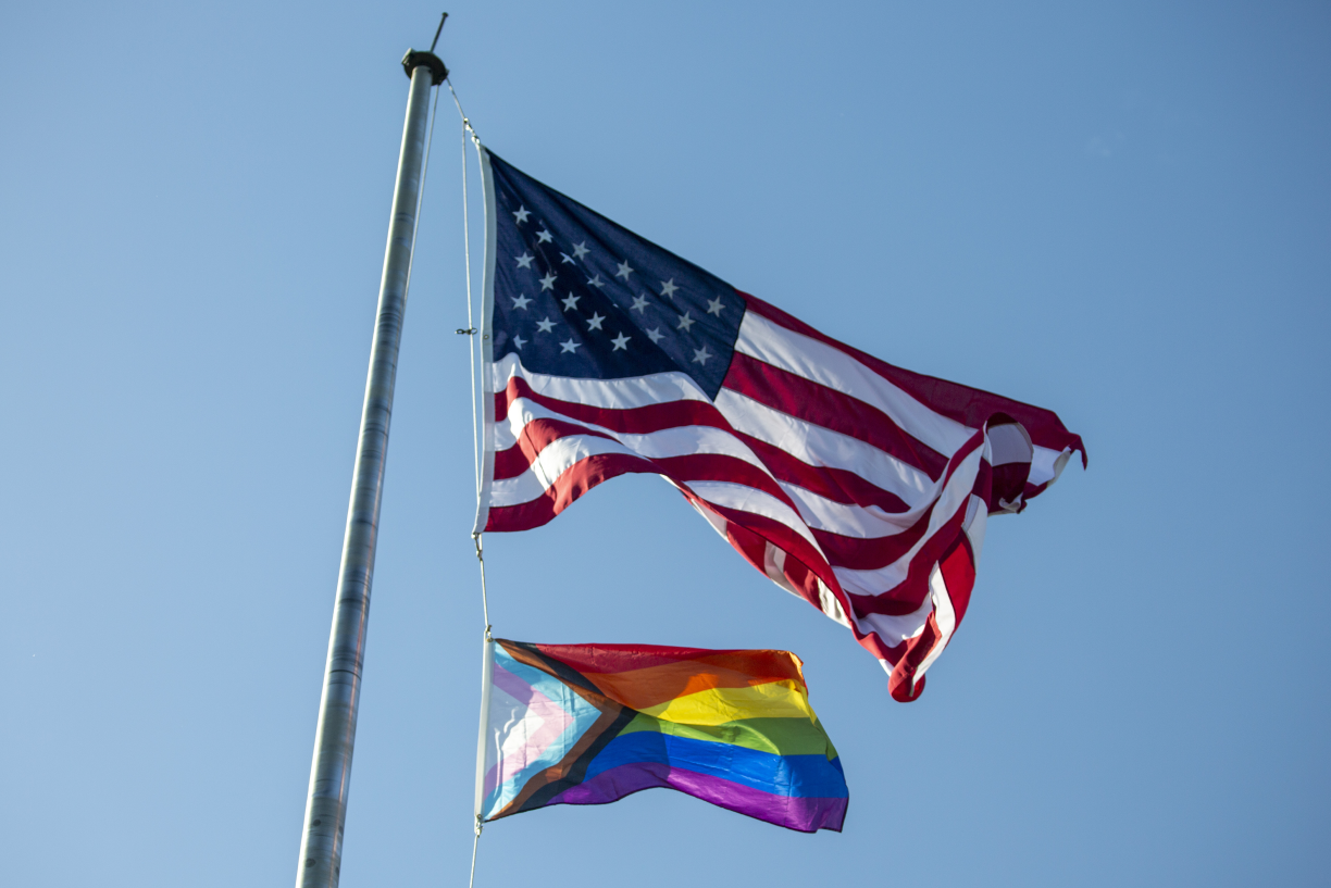 An American flag flies over the Pride Flag against a clear blue sky.