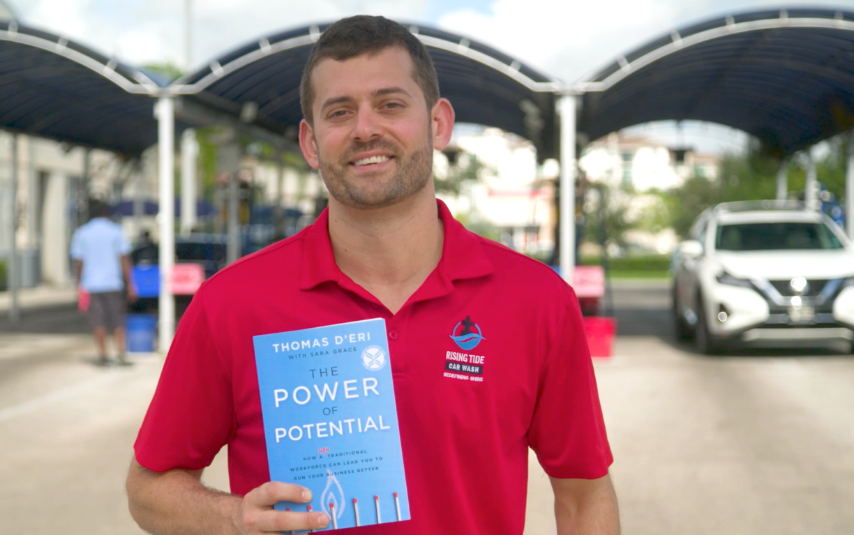 Bentley alumnus Tom D'Eri ’11 holds a copy of his new book, "The Power of Potential." In the background, employees of Rising Tide Car Wash clean several vehicles.