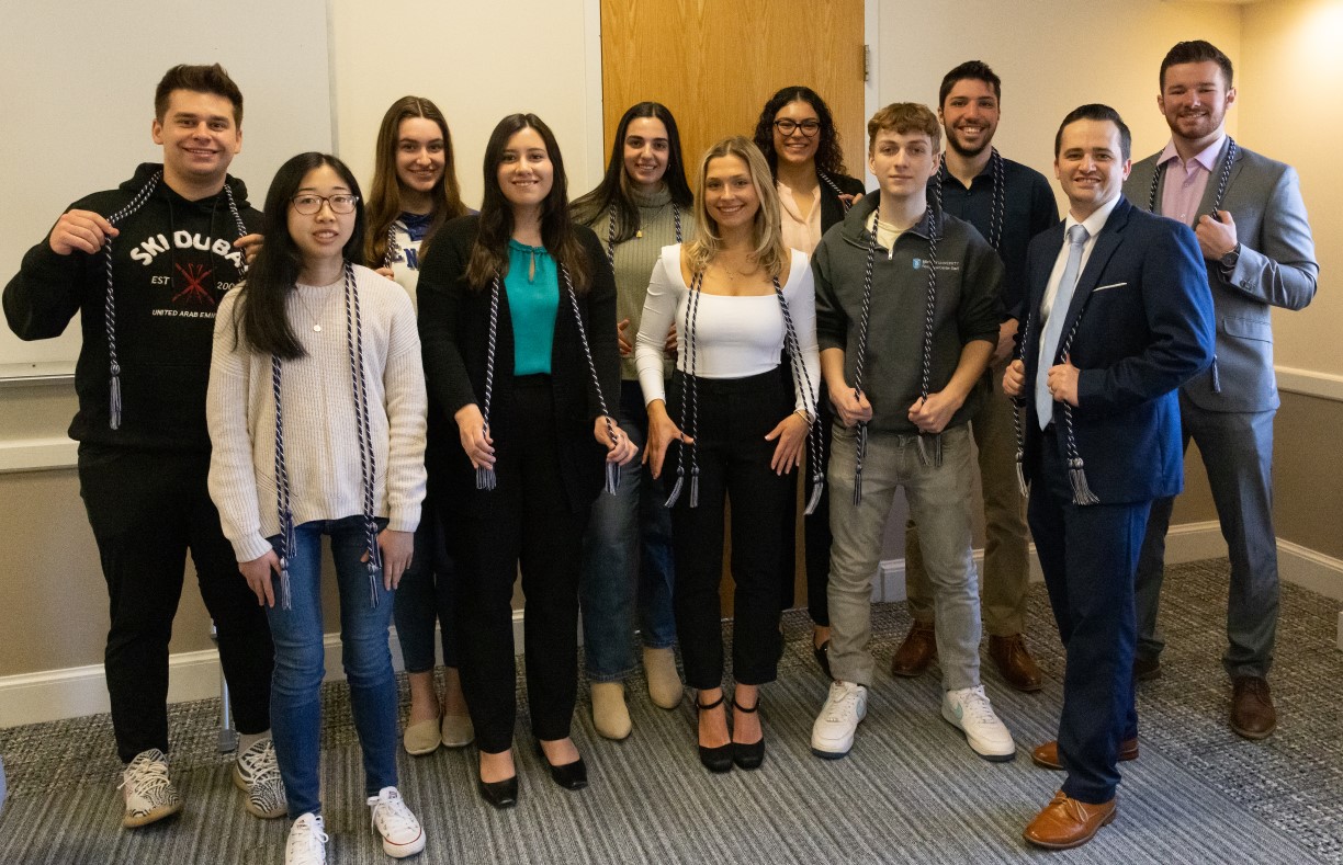 Group photo of 11 members of Bentley's Class of 2023 who were inducted into the inaugural class of Tri-Alpha. All wear tasseled cords in blue and gray —the honor society's colors — that they will wear on graduation day to denote their membership.
