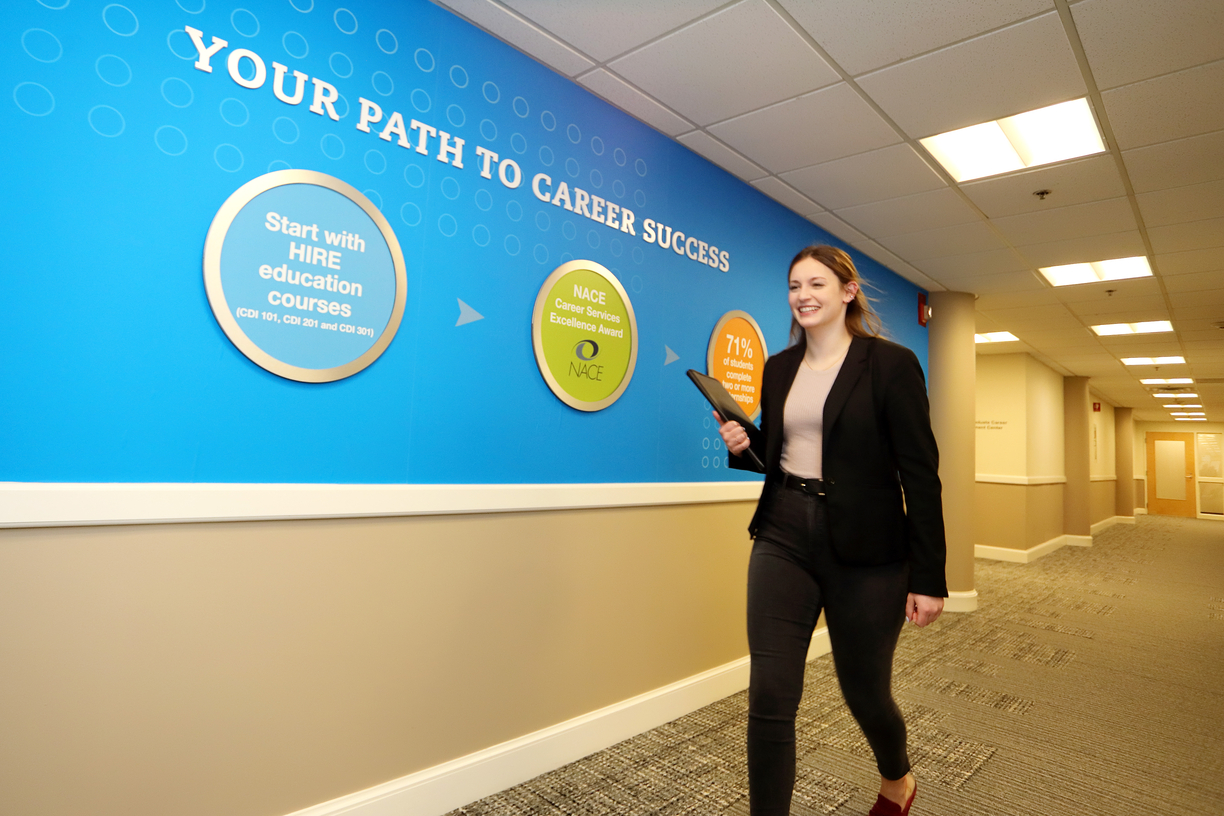 Woman in business-casual attire walking down a hallway headed toward the Bentley University Pulsifier Career Center. Bentley branded signage on the wall states: "Your Path to Career Success"