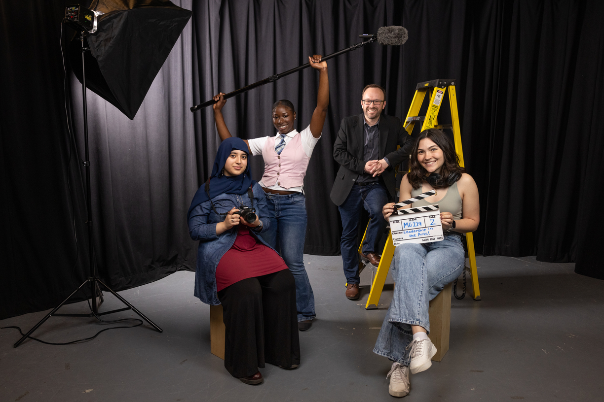 Three Bentley students and their professor pose with props — including a SLR camera, boom mike and film clapper — that illustrate the theme of their course: Leadership in the Arts.