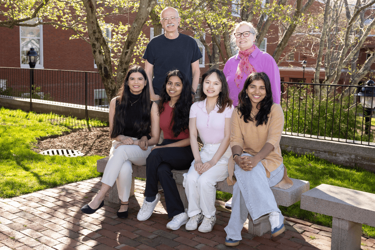 Group photo featuring four female MSHFID students seated on a stone bench, with two Bentley university professors standing behind them.