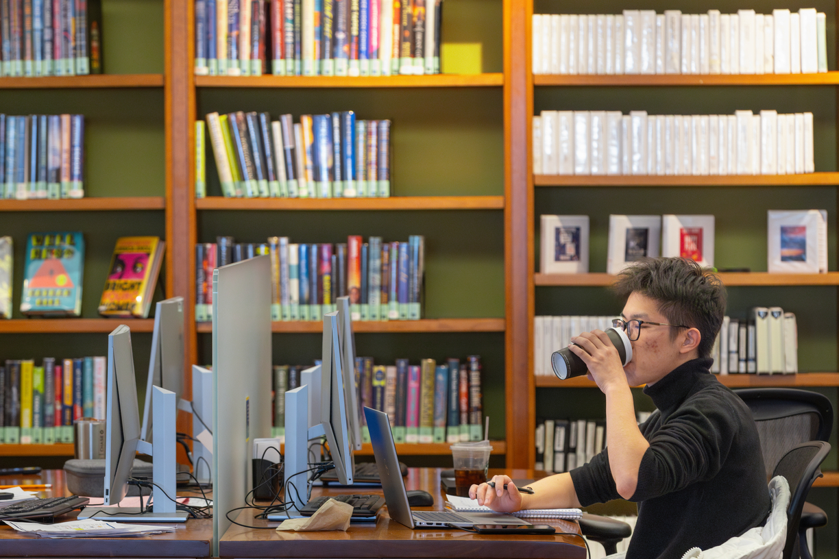 A Bentley student drinks coffee while working on a laptop in front of a bookshelf lined with colorful books