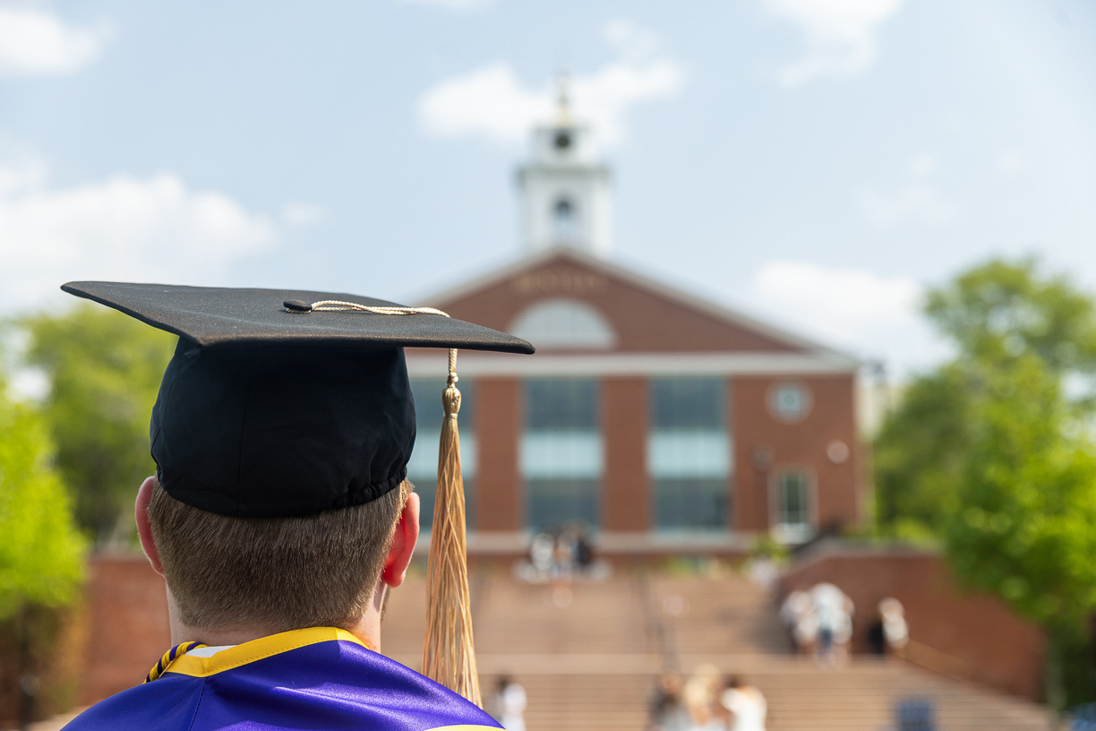 Back of a Bentley college student wearing graduation cap looking at the library clock tower as if reflecting on the future