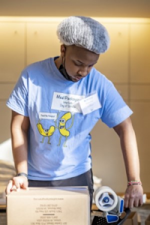 A male student uses packaging tape to seal a cardboard box of prepared meals for members of the Waltham community