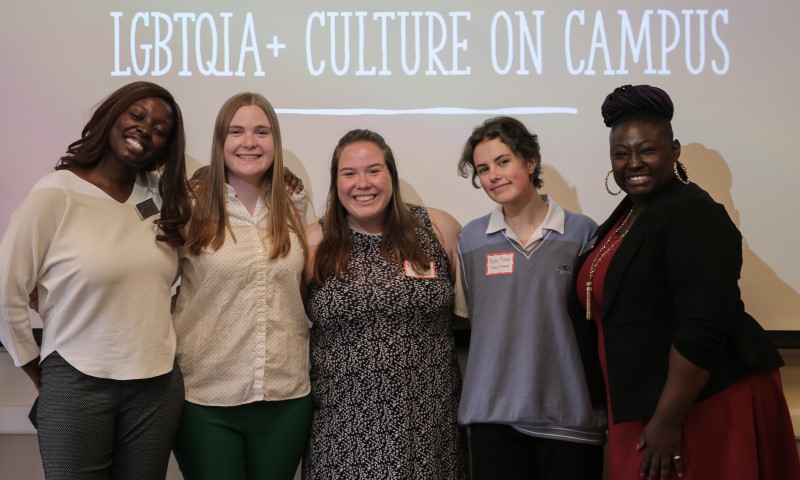Three Rainbow Scholars pose with their program leaders in front of a projection screen with the words "LGBTQIA+ Culture on Campus"