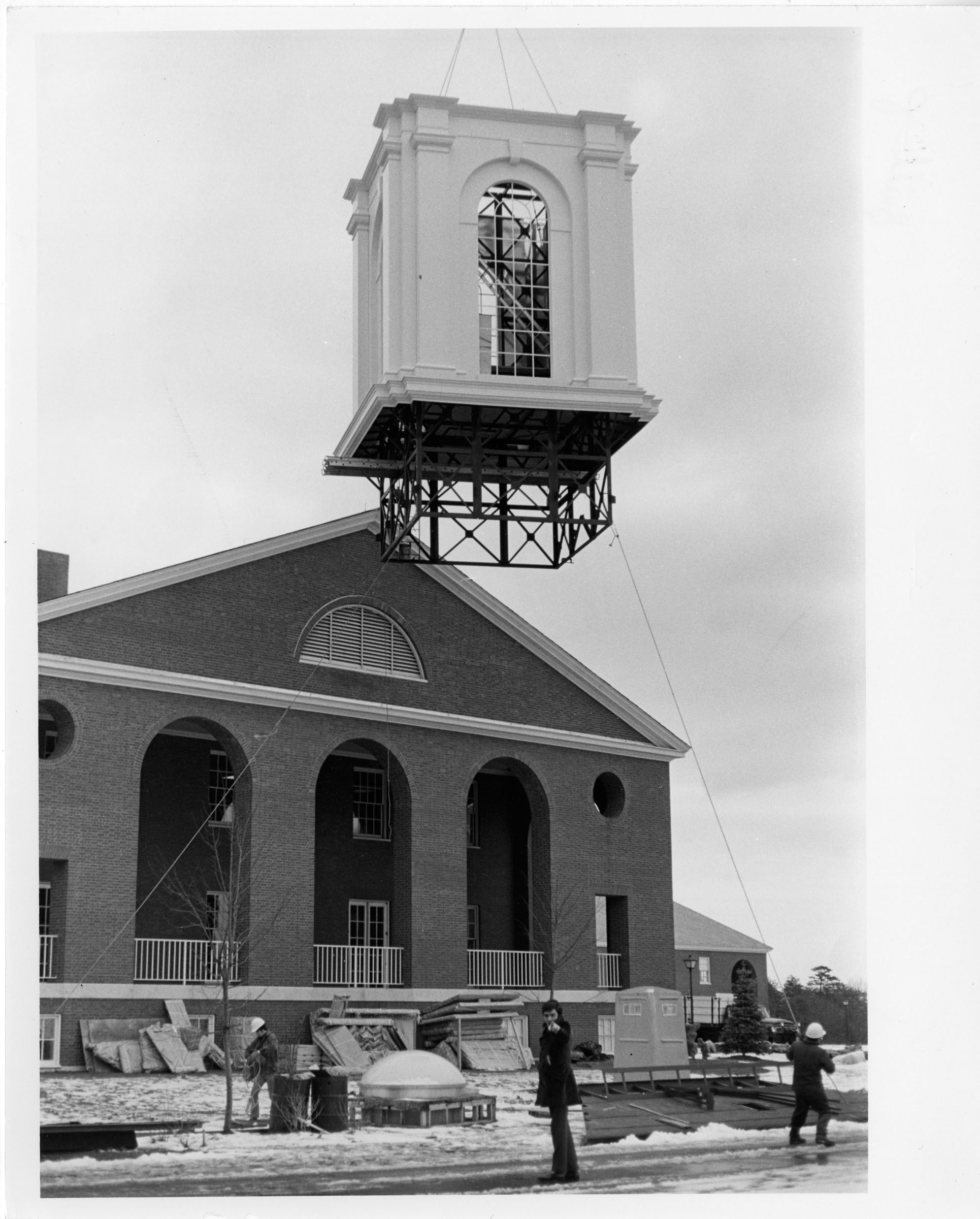 One part of the Library clocktower hangs in the air as it's being lifted to be placed. A man in the foreground points at the camera.