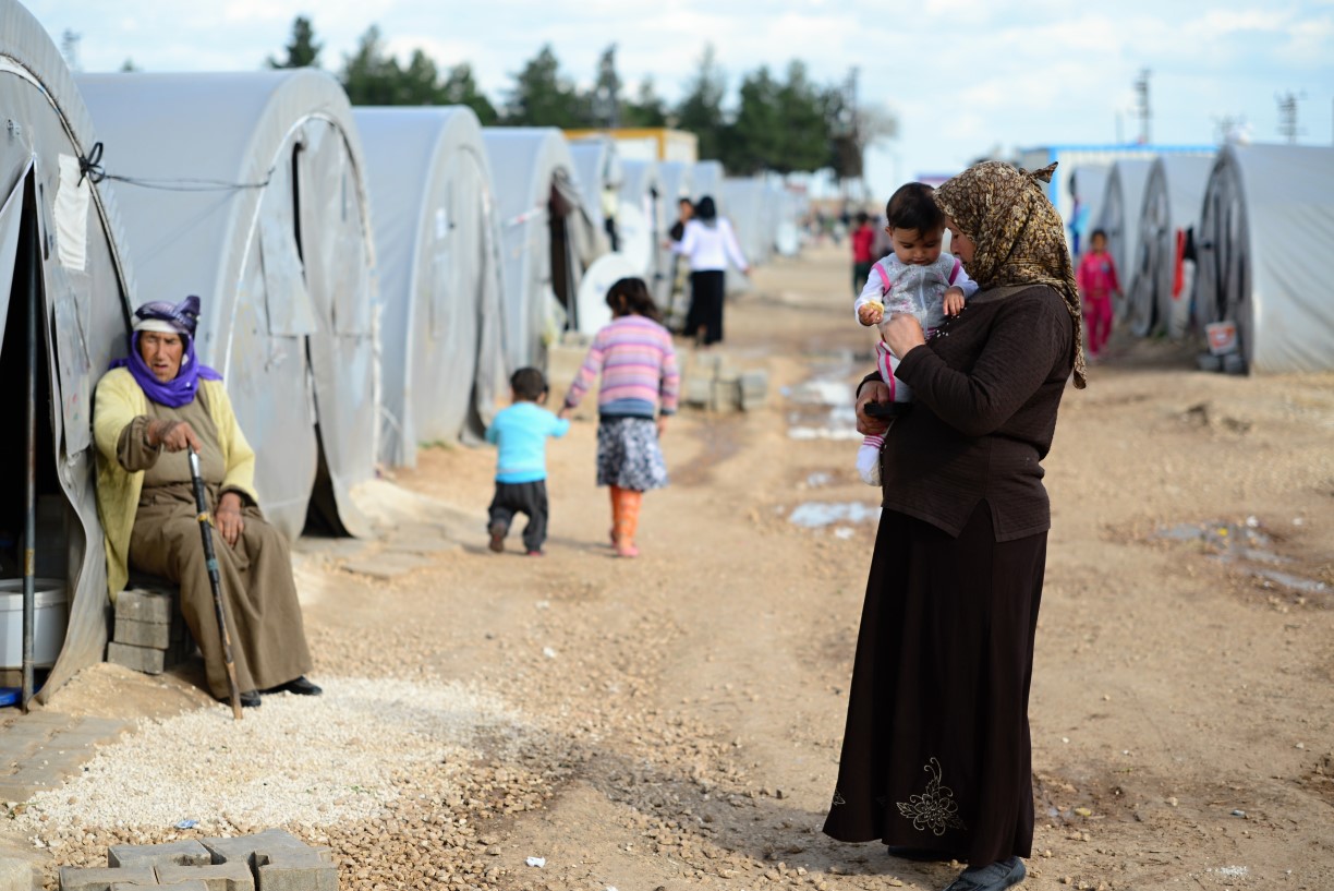 An older Syrian woman wearing a headscarf, long-sleeved shirt and ankle-length skirt cuddles a toddler as she stands in the middle of a dirt pathway within a tented refugee camp.