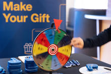 A hand spins a wheel in front of a sign that reads Make Your Gift