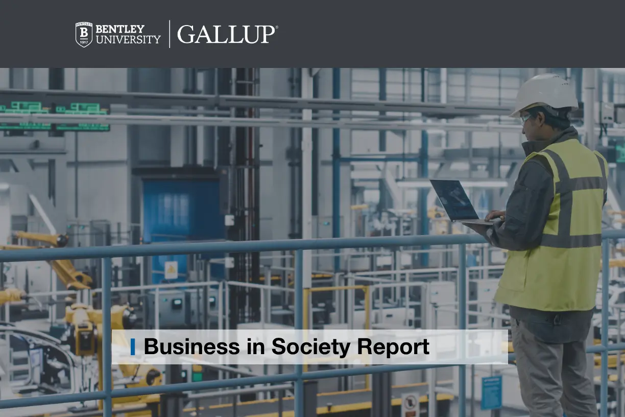 A male engineer in a yellow jacket and construction hard hat holds an open laptop while surveying an auto manufacturing plant