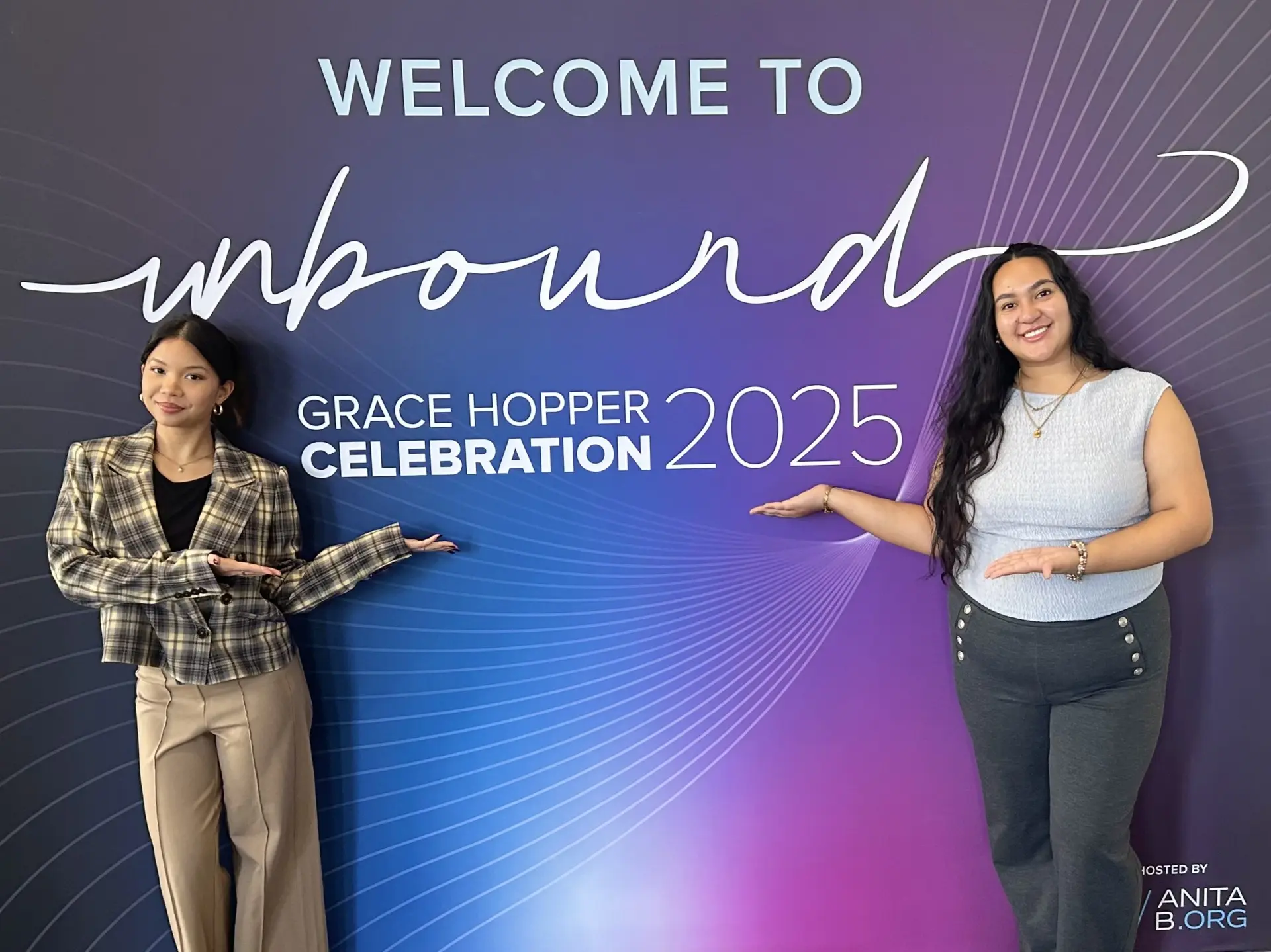Two Bentley University female students stand in front of a blue and purple big sign that reads "Welcome to Unbound: Grace Hopper Celebration 2025" 