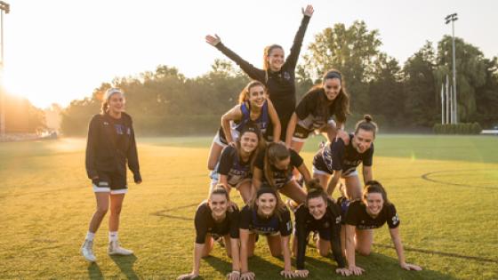 women smile on lacrosse field while making human pyramid