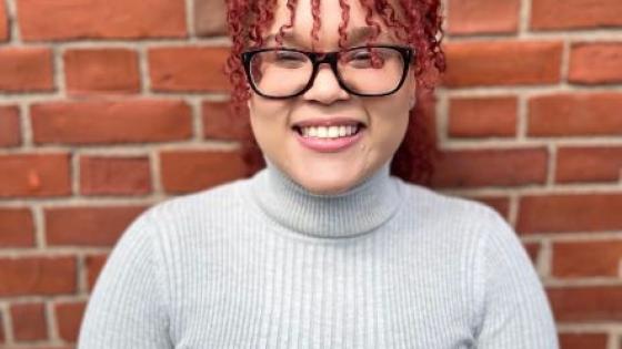 Nicole standing in front of a brick wall in a gray turtleneck, black square glasses, and smiling