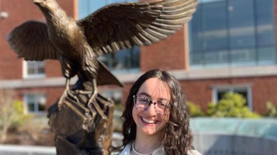 Yasmin standing in front of the bronze falcon statue wearing a white long sleeve blouse, glasses and smiling