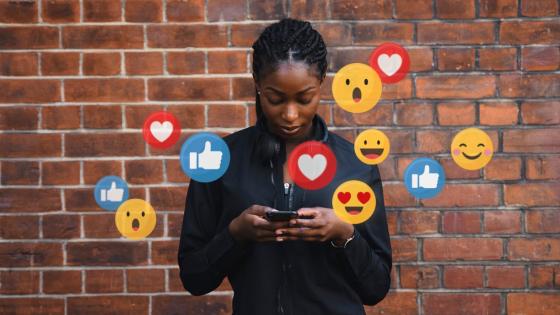 A young Black woman stands in front of a brick wall, staring down at her smartphone. A dozen different social media icons and emojis — including hearts, thumbs-up "likes" and smiley faces — surround her.