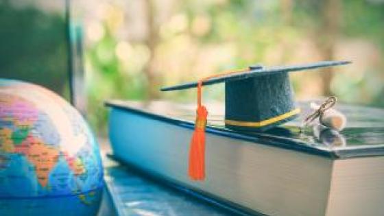 A graduation cap sitting atop a texbook next to a globe. 