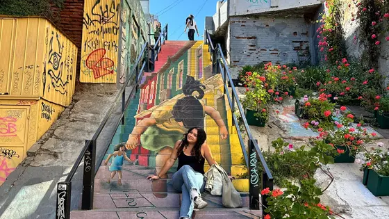 A vibrant staircase mural in Valparaiso, Chile, depicting a person carrying a child on their back with buildings and streets in the background. The artwork creates a sense of depth and perspective across the steps. Graffiti decorates the adjacent walls, and blooming flowers in planters line the right side of the staircase. A person is seated on one of the steps, posing for a photo, adding a human element to the colorful urban scene.
