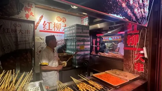  A vibrant street food vendor stall at an Asian market, specializing in skewered meat. Various skewers are being grilled and displayed in trays at the front of the stall. A large container of seasoning or sauce sits nearby. Bright signage with Chinese characters decorates the stall, and several people are visible behind the counter preparing and serving food.