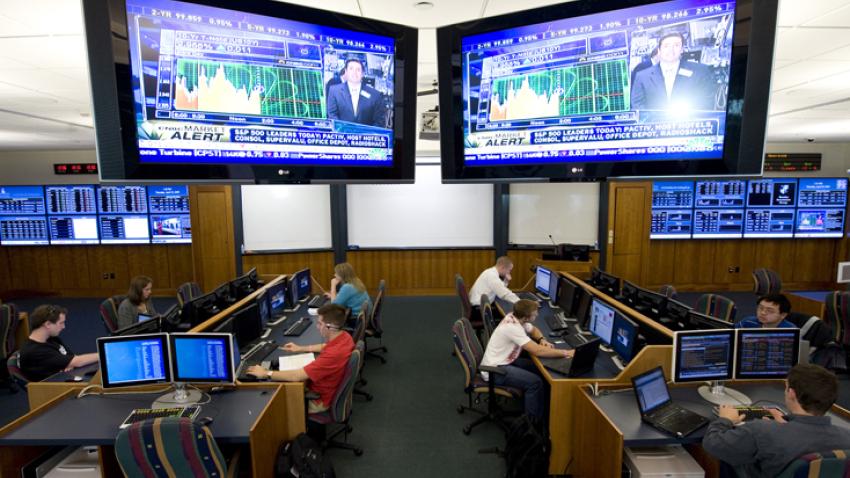 Students working on computers in a large room with two large monitor screens in the front of the room. 