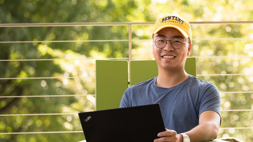 Student on laptop outside wearing a Bentley University hat