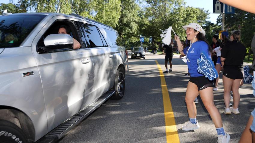 Bentley orientation leader cheers as incoming students arrive and pass in cars