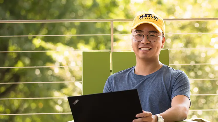 Student on laptop outside wearing a Bentley University hat
