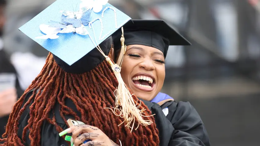Graduates embrace at the 2022 Commencement ceremony. One woman's cap is adorned with blue butterflies and says "22" in beading across the top. 
