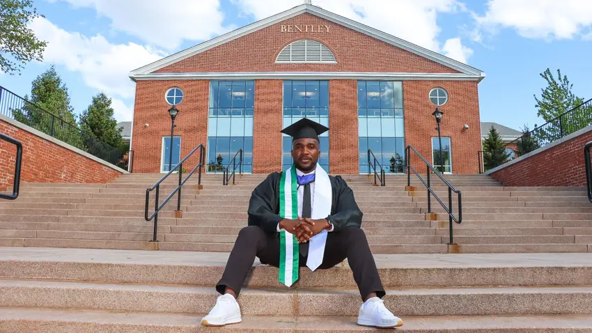 Victor Obetta sitting on steps in front wearing graduation robe and cap