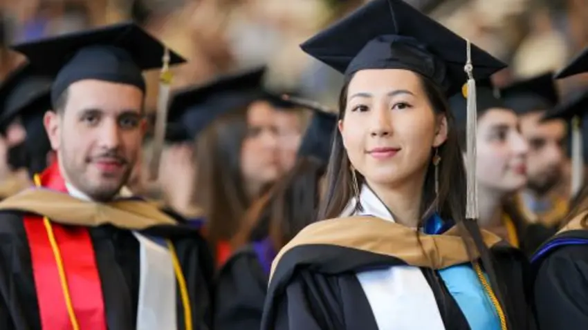 A Class of 2023 graduate wearing commencement regalia sitting with fellow graduates