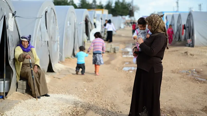 An older Syrian woman wearing a headscarf, long-sleeved shirt and ankle-length skirt cuddles a toddler as she stands in the middle of a dirt pathway within a tented refugee camp.