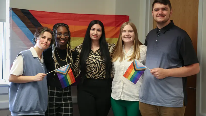 Five members of the inaugural class of Rainbow Scholars pose in front of a rainbow flag. Two also hold smaller rainbow flags in their hands.