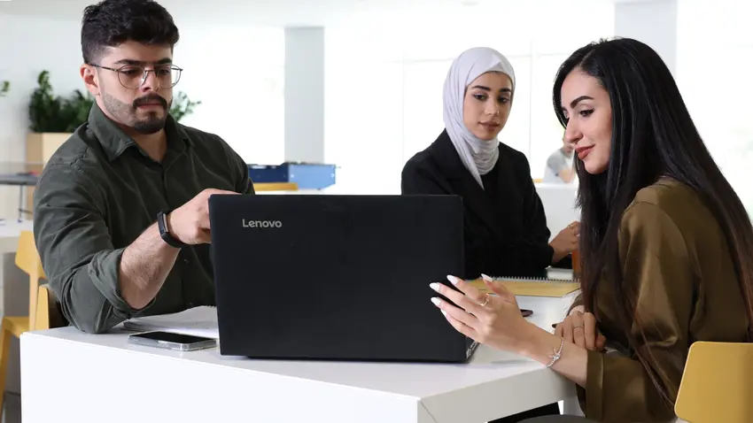 Three Kuwait College of Science and Technology students, one male and two female, sit together at a white table and look toward an open laptop computer.
