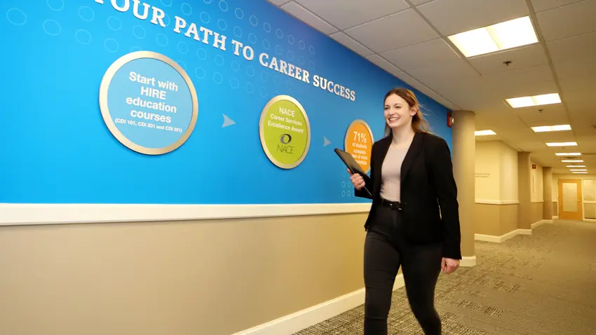 Woman in business-casual attire walking down a hallway headed toward the Bentley University Pulsifier Career Center. Bentley branded signage on the wall states: "Your Path to Career Success"