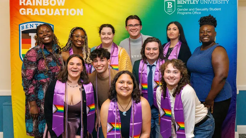 11 individuals pictured smiling, wearing graduation stoles and cords against a colorful Rainbow Graduation backdrop