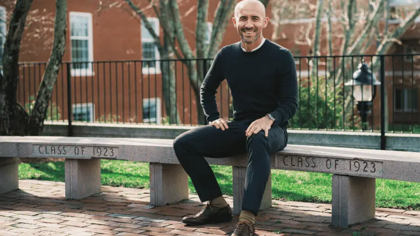Economics professor sits on a curved concrete bench engraved with "Class of 1923."