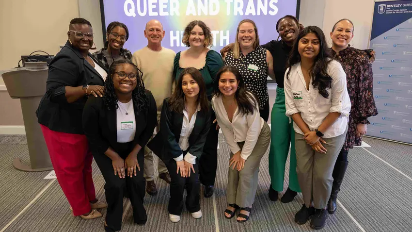 Image of 11 individuals smiling and posing in front of a "Queer and Trans Health" presentation slide