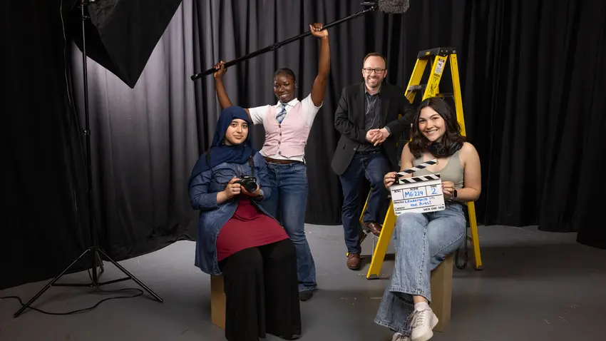 Three Bentley students and their professor pose with props — including a SLR camera, boom mike and film clapper — that illustrate the theme of their course: Leadership in the Arts.