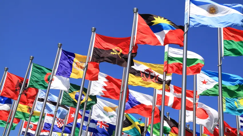 Dozens of flagpoles bearing the flags of different countries waving in the breeze against a backdrop of blue sky.