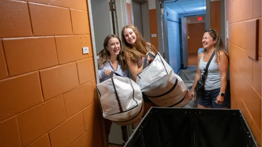 Bentley students holding large bags in dorm hallway as parent laughs during Class of 2029 move-in day