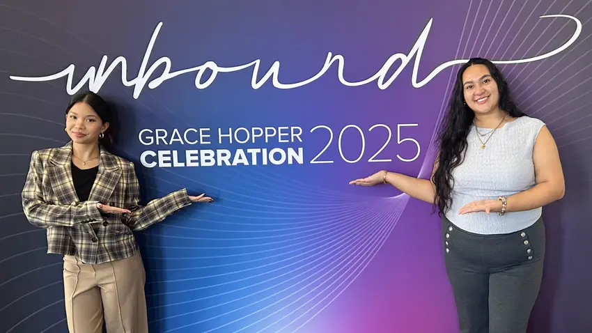 Two Bentley University female students stand in front of a blue and purple big sign that reads "Welcome to Unbound: Grace Hopper Celebration 2025" 