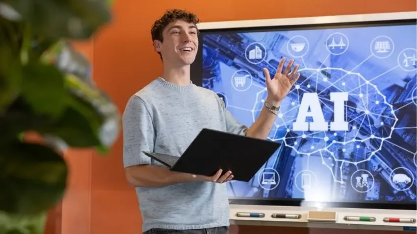 Bentley student Aidan Griffith in the CIS Sandbox, holding a laptop in front of a screen with AI imagery