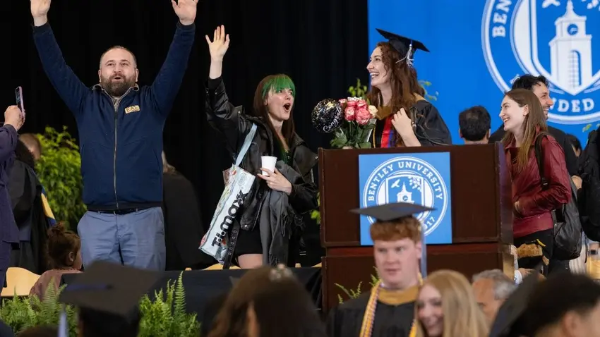 A graduate holding a bouquet of flowers celebrates with friends and family on stage after the Bentley Class of 2026 McCallum Graduate School of Business 51st commencement ceremony. 