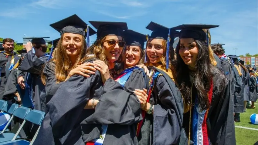 Friends from the Bentley Class of 2026 lean in to pose together in caps and gowns during Bentley commencement. 