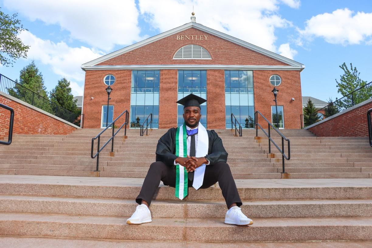 Victor Obetta sitting on steps in front wearing graduation robe and cap